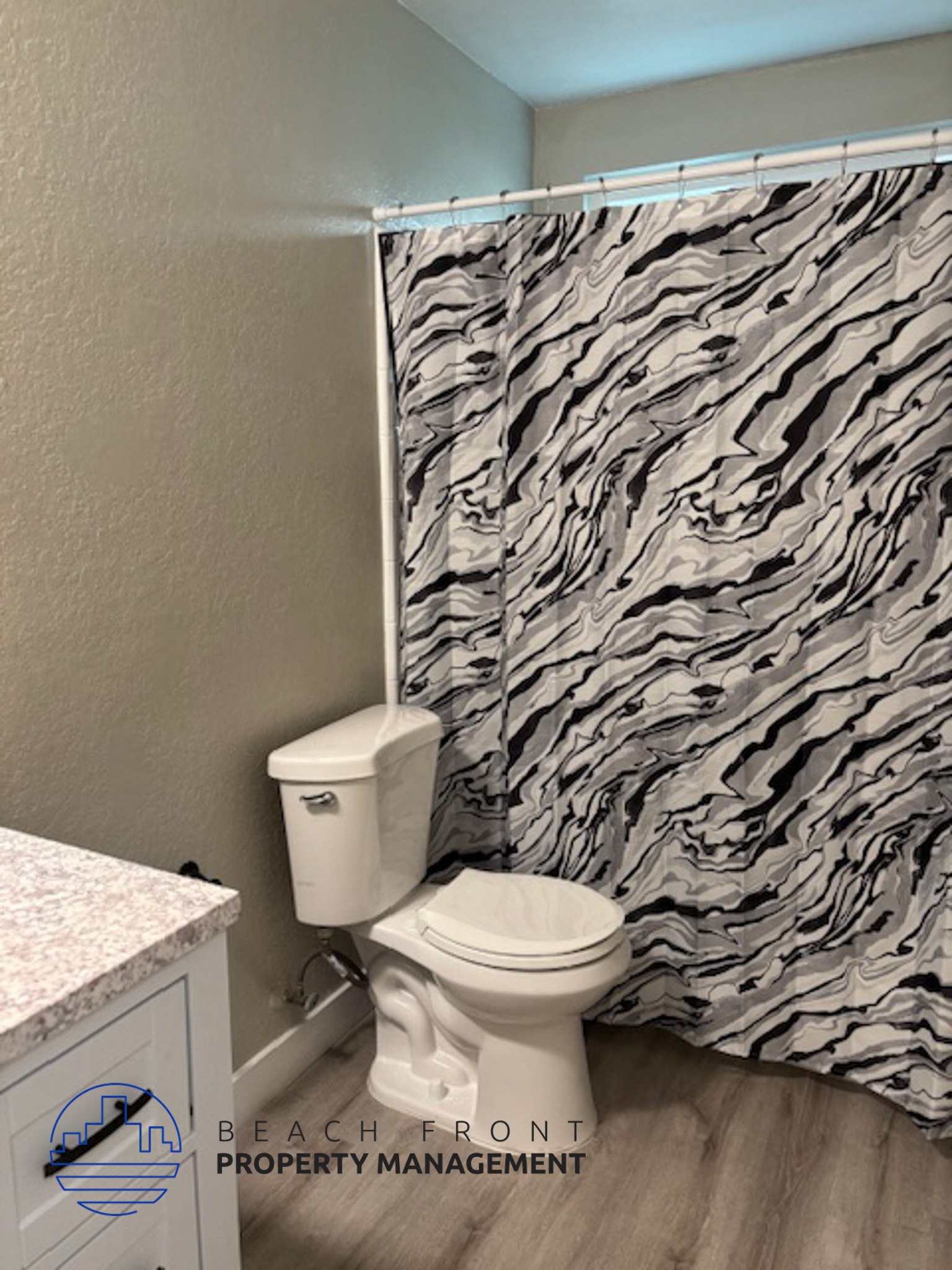 A bathroom with a toilet, a shower curtain with a marbled pattern, and a cabinet with a logo for Beach Front Property Management.