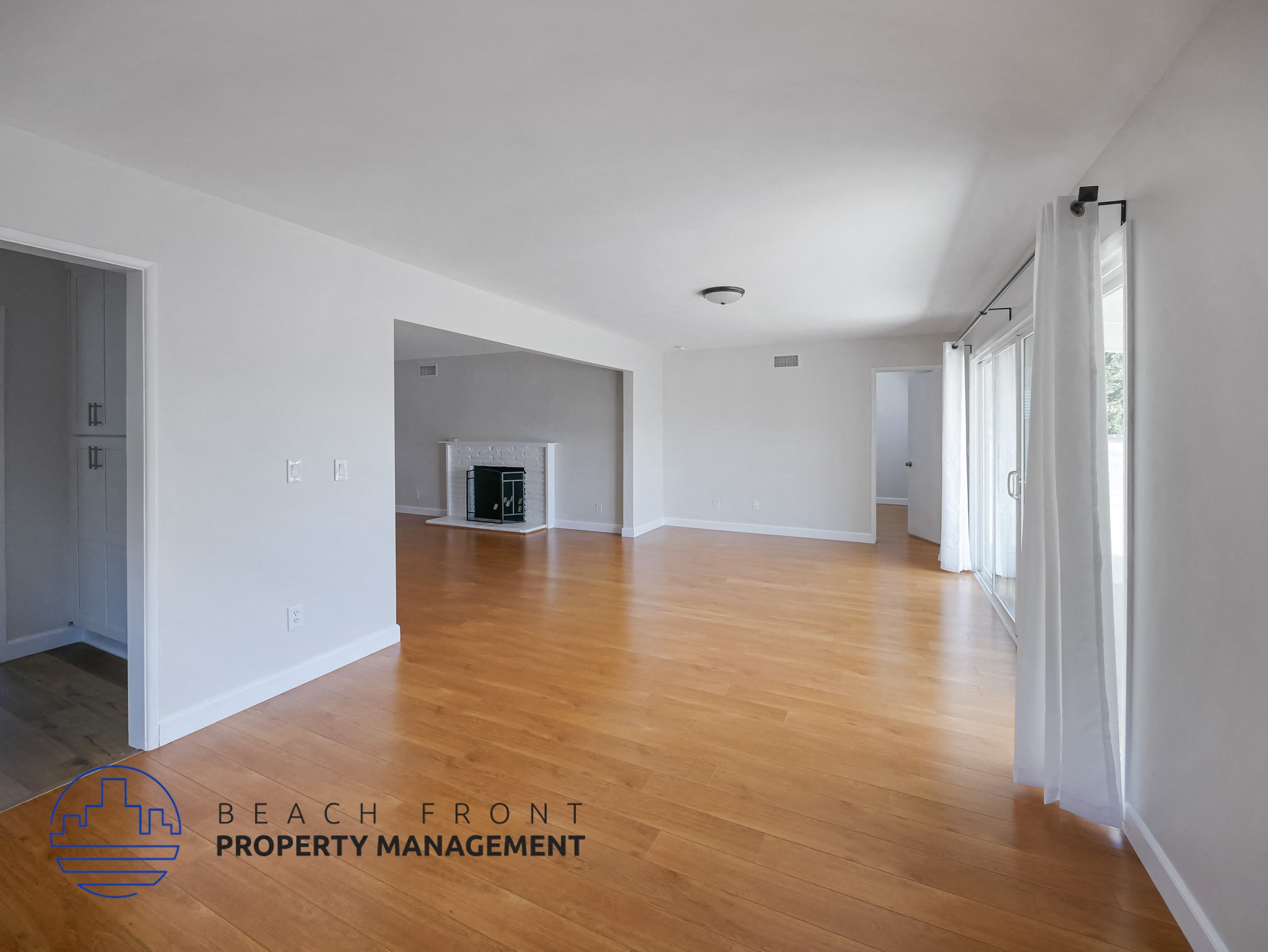 A spacious room with wooden floors and white walls, with a logo for Beach Front Property Management in the bottom left corner.