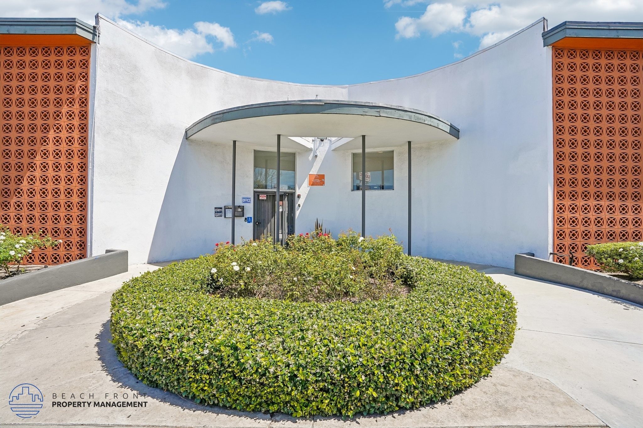 A white building with orange patterned walls and a circular green bush in front.