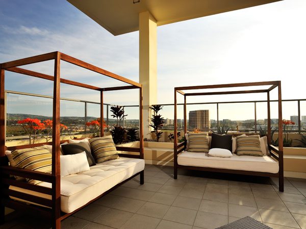 Apartments in Irvine, CA - Astoria at Central Park West - Outdoor Patio With Two Wooden Daybeds, Cushioned Seating, Glass Railings, Planters, and Expansive City Views at Sunset.