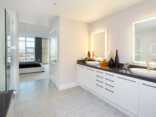 Irvine, CA, Apartments - Astoria at Central Park West - Contemporary Bathroom With Double Vanity, Backlit Mirrors, Sleek Countertop, Glass Shower, and Light Tile Flooring Leading To Bedroom.