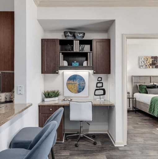 Apartment interior featuring a built-in desk with overhead cabinets, a white office chair, and a glimpse into the bedroom.