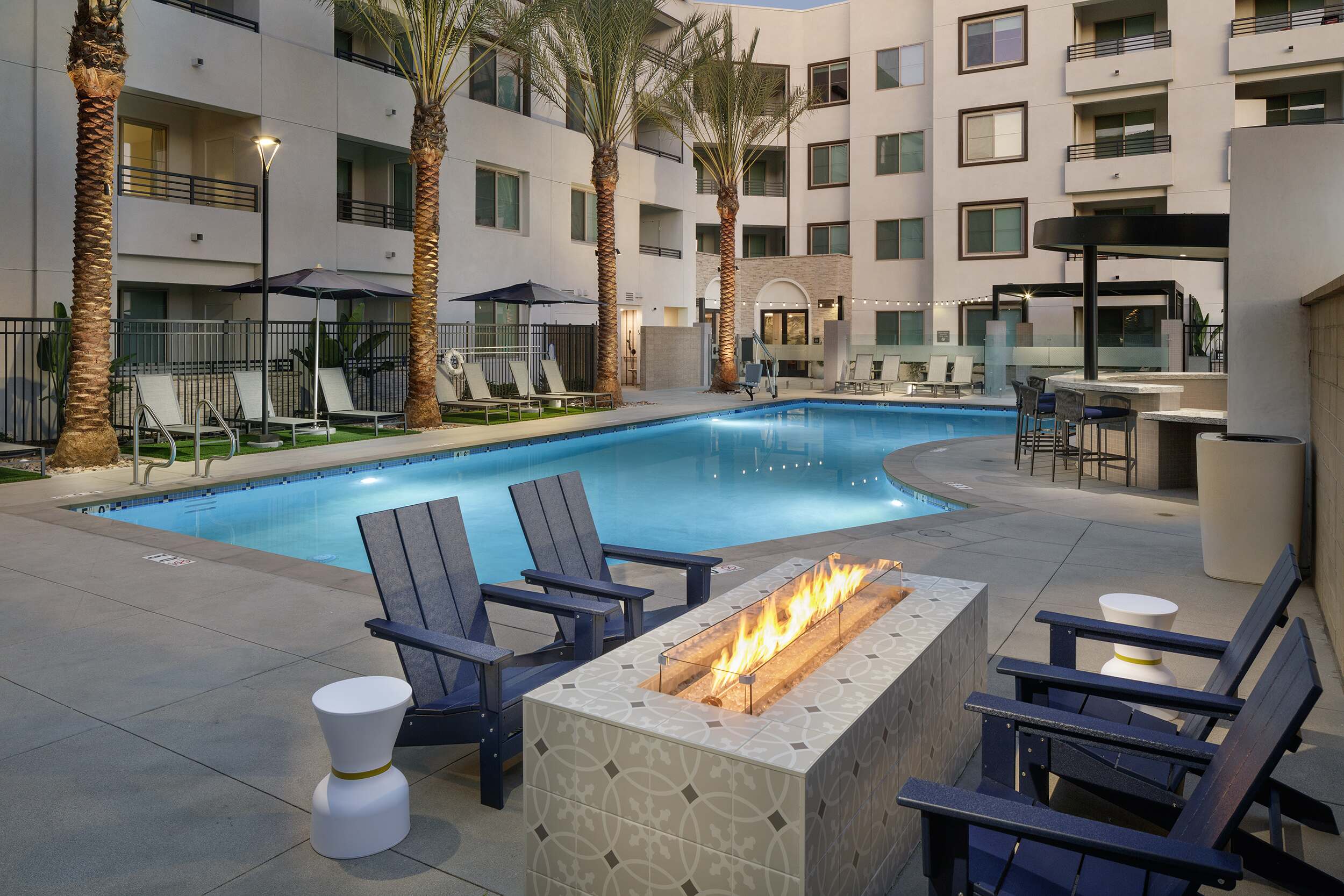 Pool courtyard at dusk with lounge chairs, palm trees, apartment balconies, and a linear fire pit.
