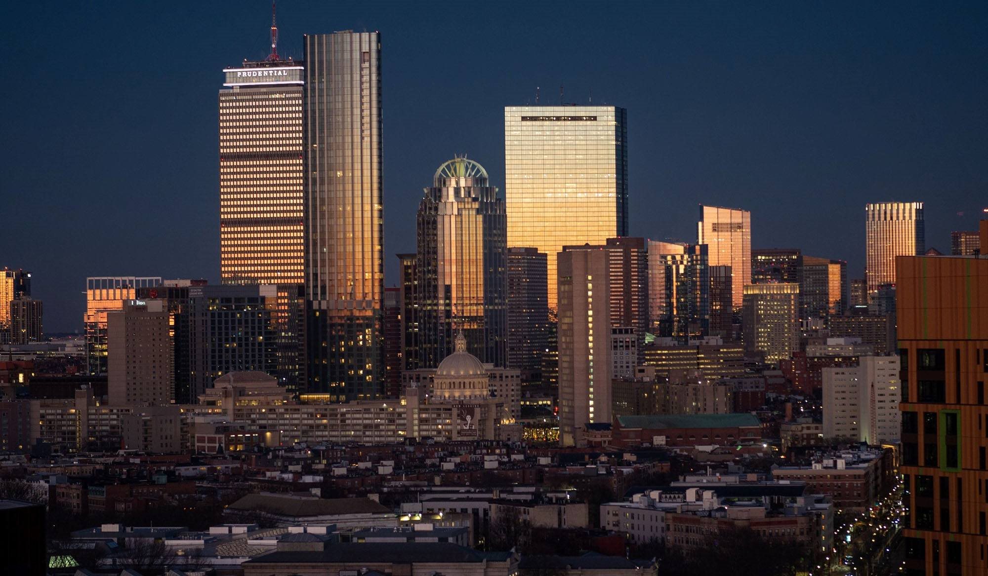 A city skyline at dusk with buildings illuminated against a darkening sky.