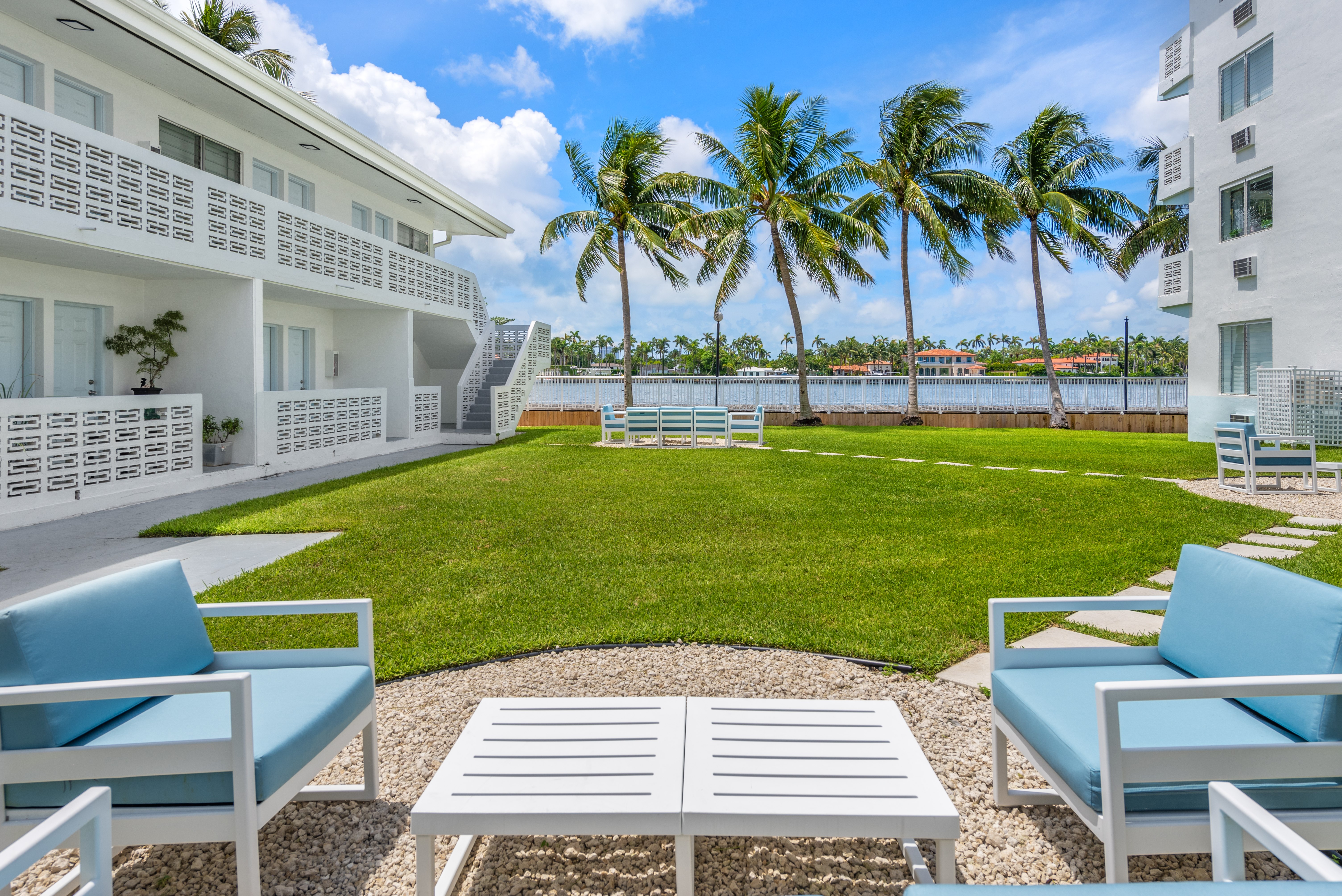 A white building with a balcony overlooks a green lawn with palm trees.