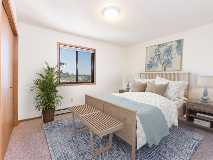 Bismarck, ND Fairview Apartments. A bedroom that showcases a comfortable bed with neatly arranged bedding, a stylish bed bench, and a window that fills the space with natural light. It's a serene and inviting setting for rest and relaxation.