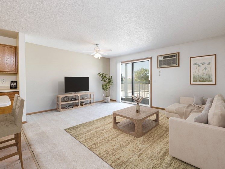 Bismarck, ND Sierra Ridge Apartments a living room with white walls and a sliding glass door. Theres a white couch, coffee table, tv and bar stools at the counter in the dining area.