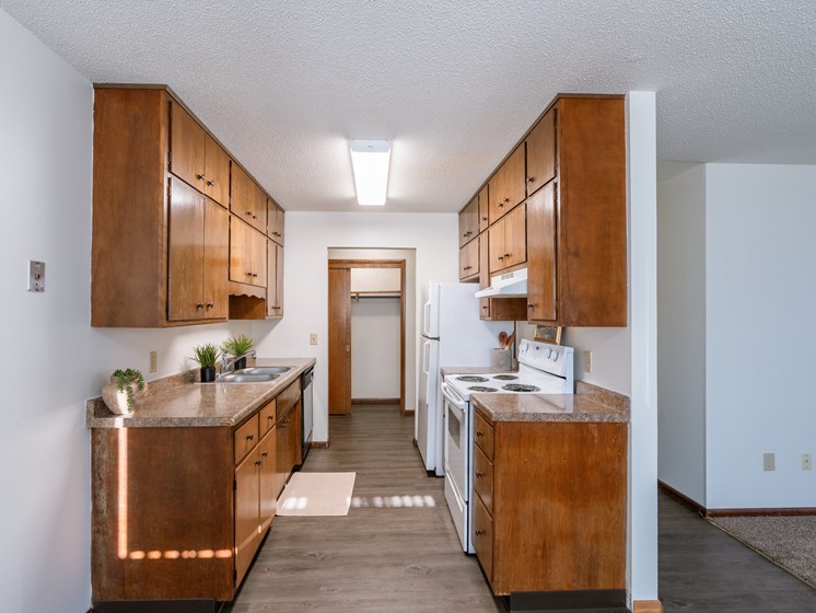 an open kitchen with wood cabinets and white appliances and a hallway with a door to. Fargo, ND Martha Alice Apartments