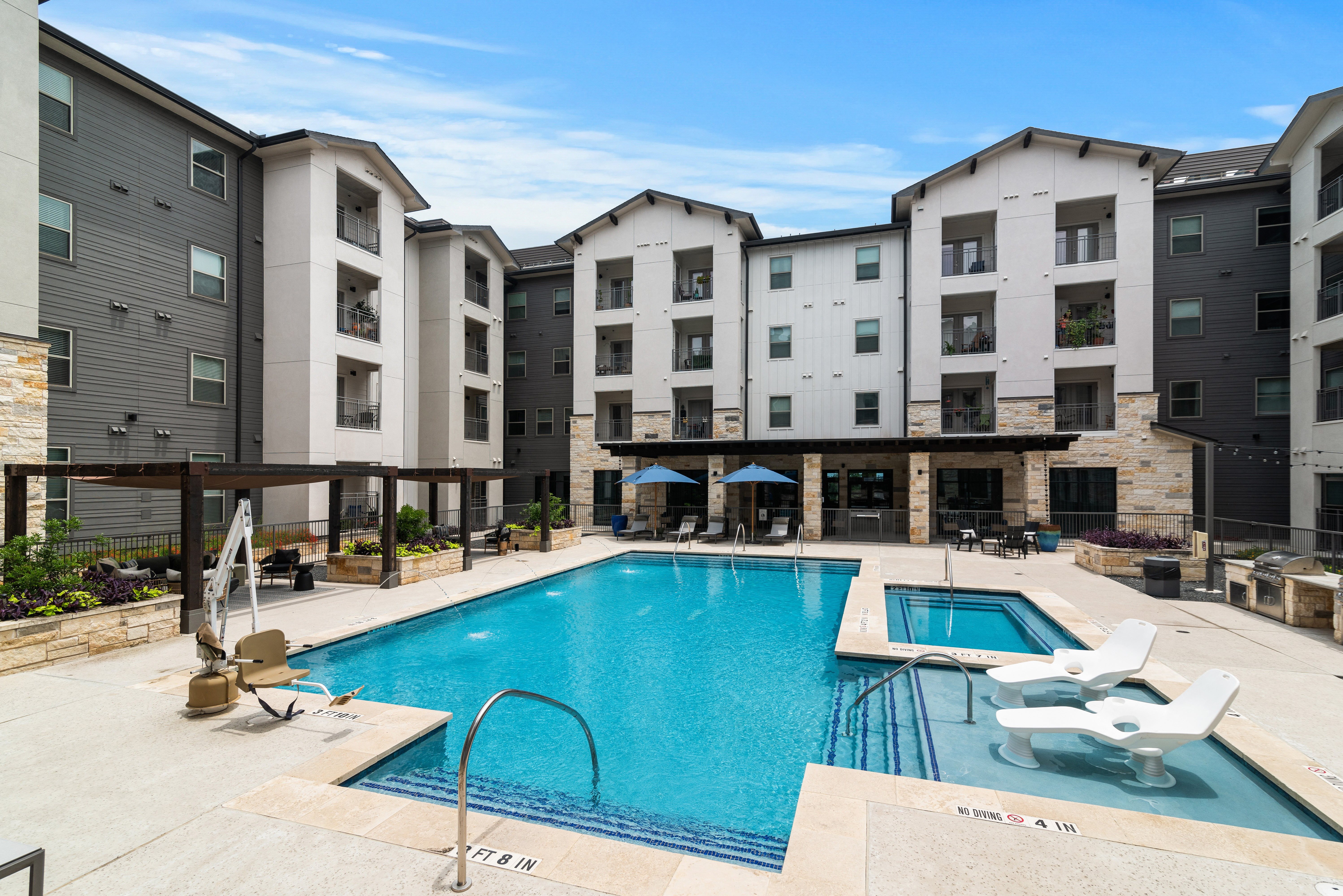 a resort-style swimming pool with lounge chairs and sitting areas covered by a wooden pergola at a senior apartment community