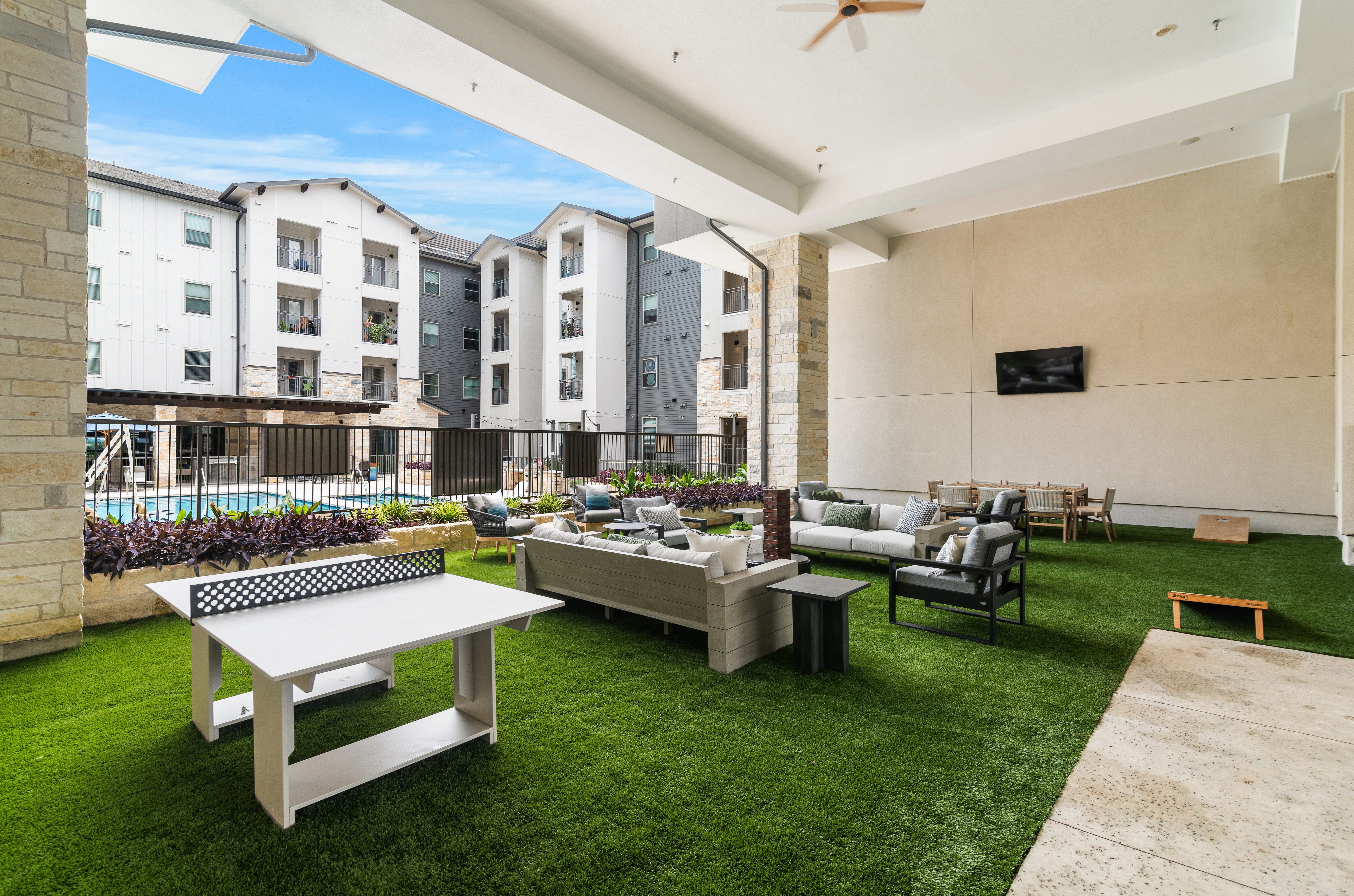 an outdoor lounge area on turf with couches, cornhole, ping pong and a flat screen tv looking at the pool at a senior apartment community
