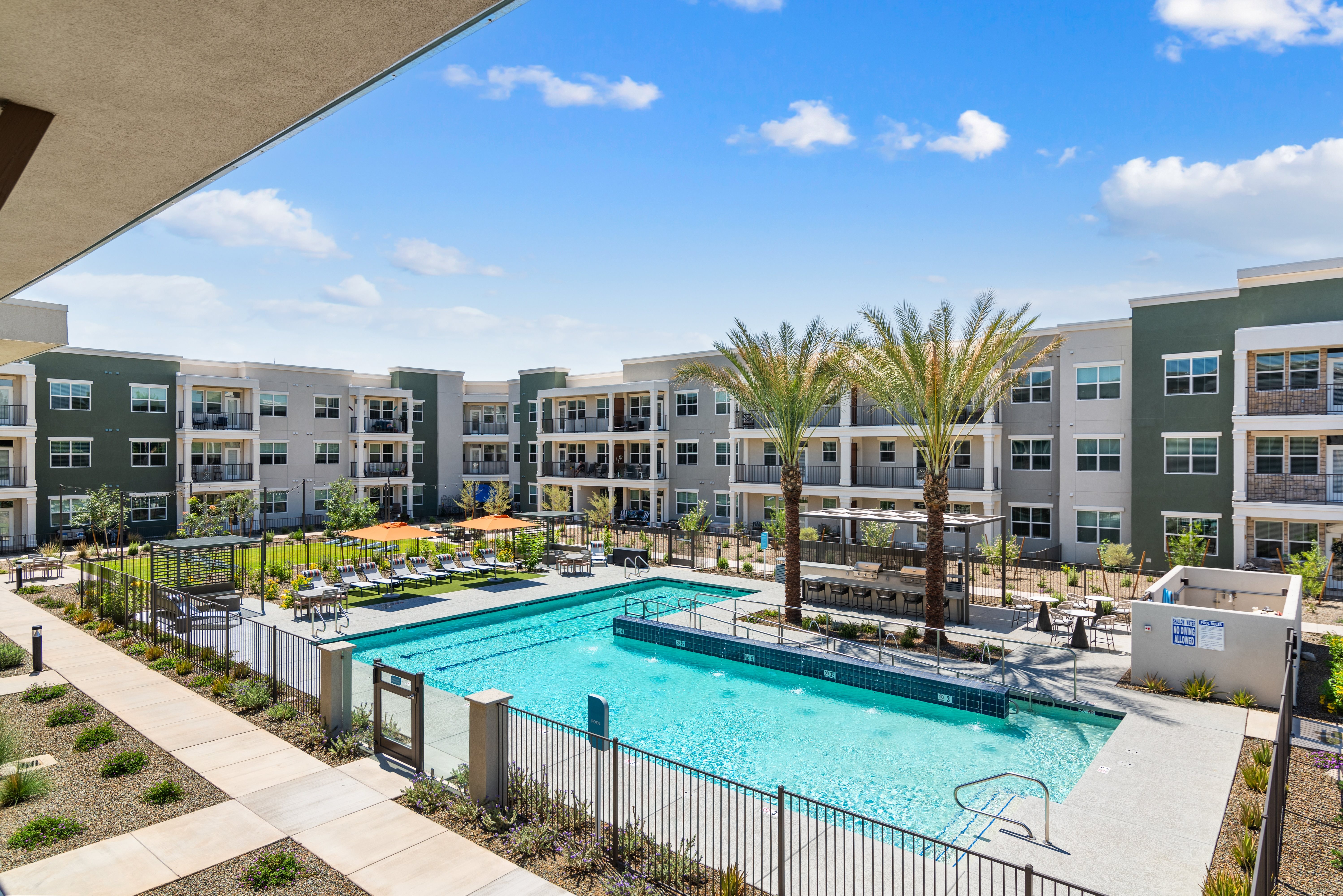 A swimming pool is surrounded by a fence and palm trees in front of apartment buildings.