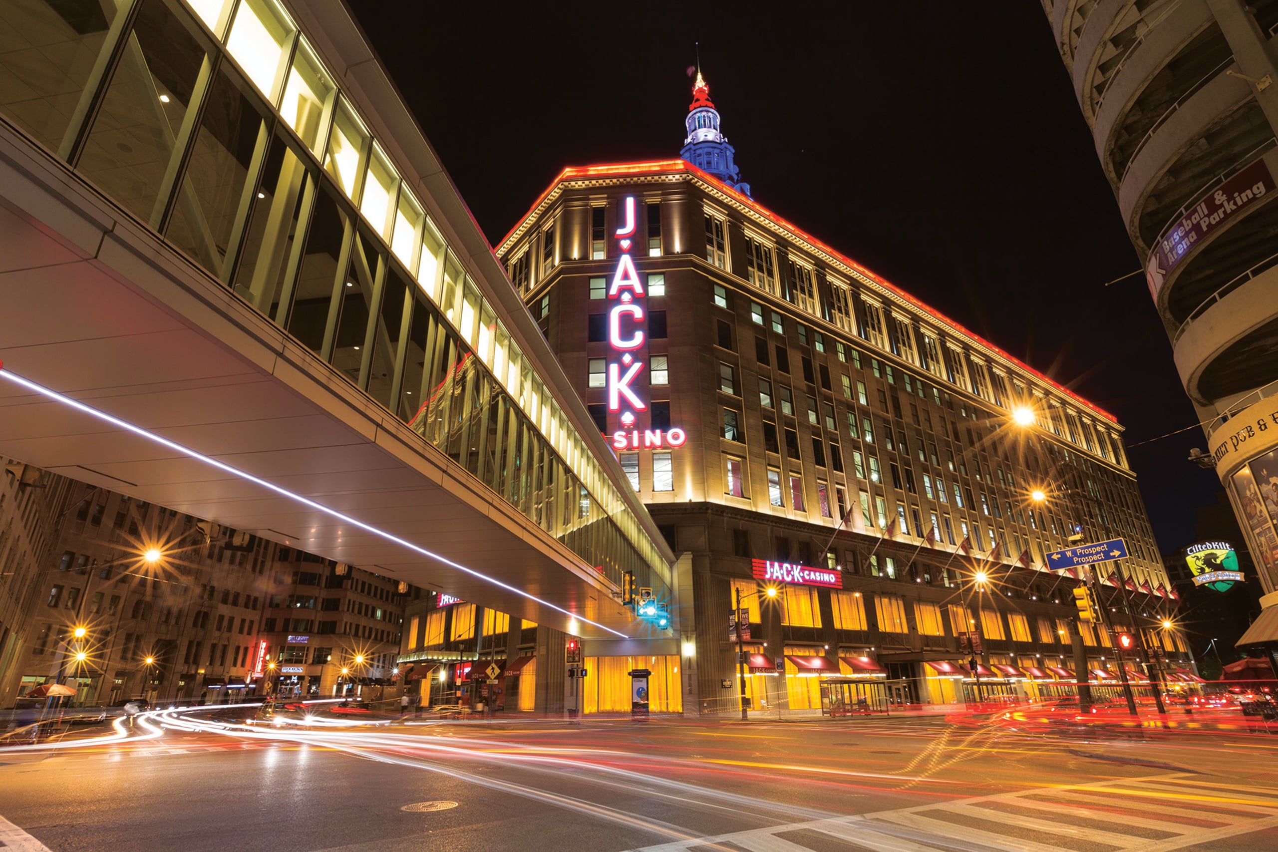 Nighttime view of the neon-lit JACK Cleveland Casino and skywalk at Public Square in downtown Cleveland, with the glowing spire of The Terminal Tower Residences rising in the background.