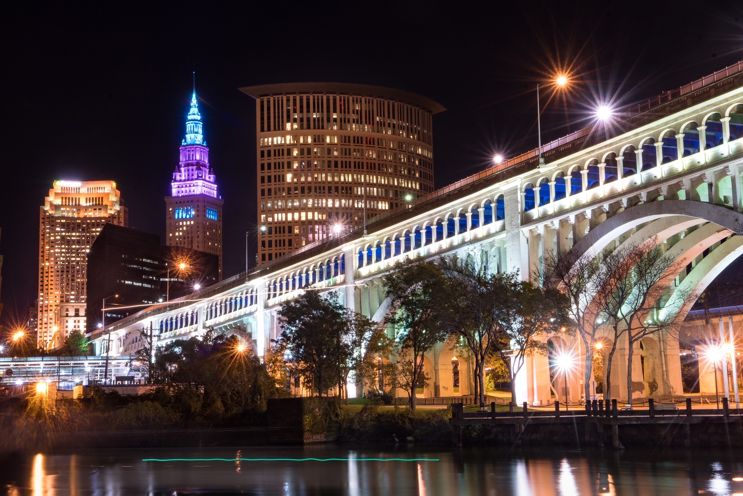 A stunning night view of the illuminated Detroit-Superior Bridge stretching over the Cuyahoga River, with the glowing blue and white lights of The Terminal Tower Residences standing tall in the downtown Cleveland skyline.