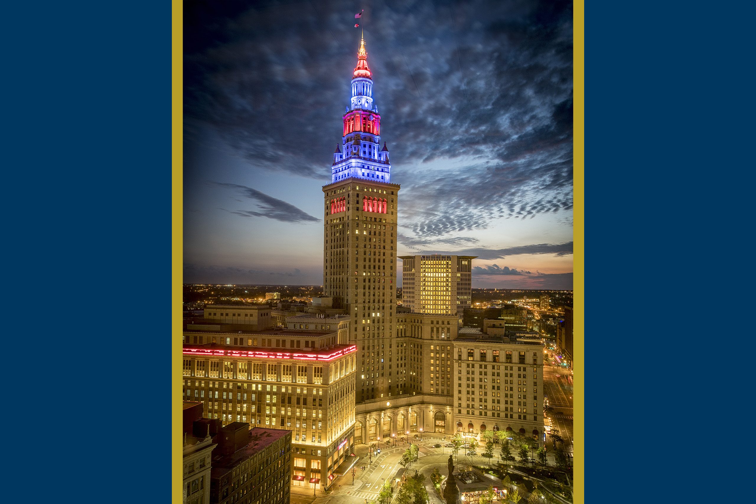 High-angle twilight view of The Terminal Tower Residences illuminated in patriotic red, white, and blue lights, soaring above the cityscape of downtown Cleveland.