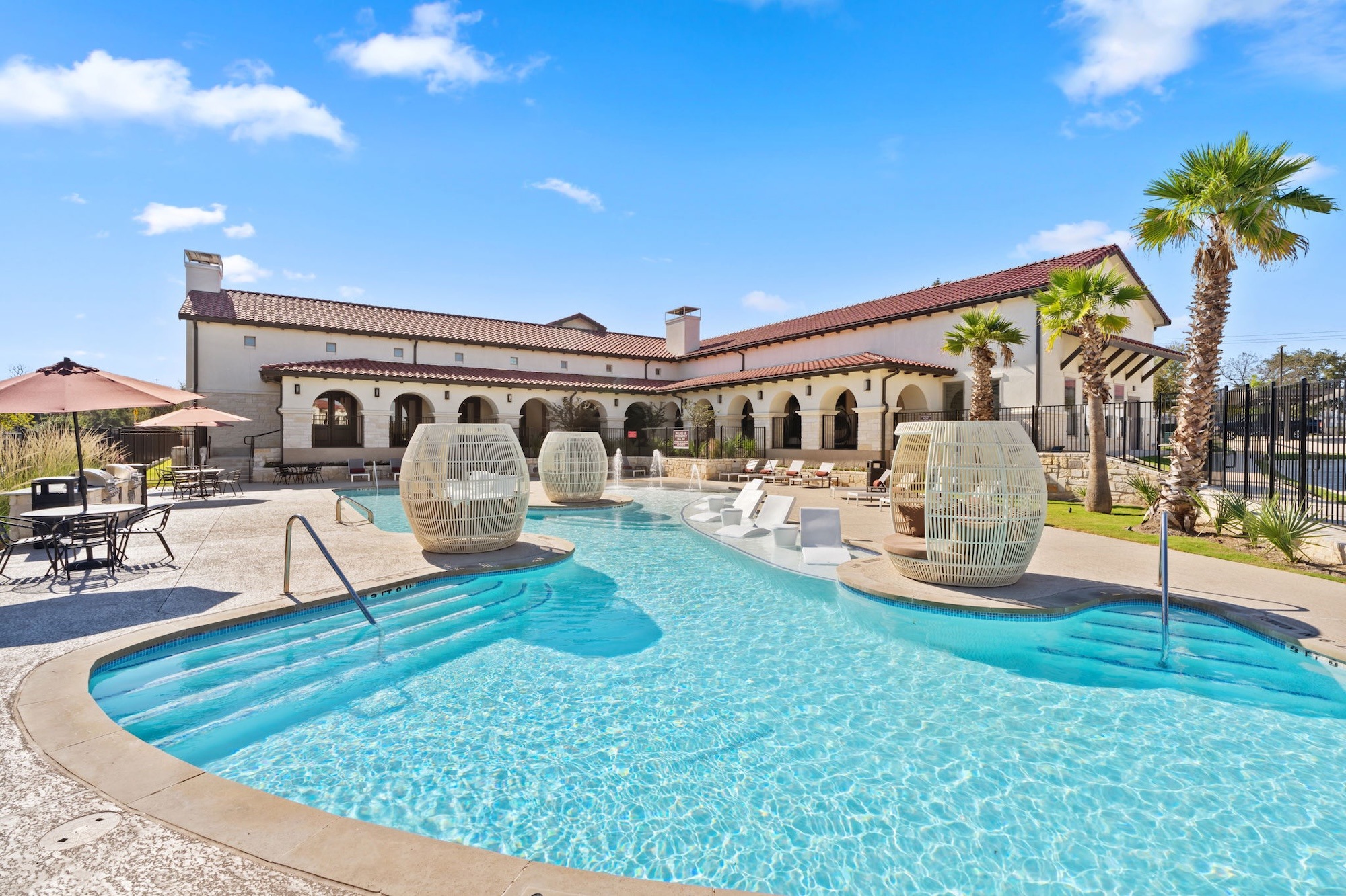 Sunny resort courtyard with turquoise pool, lounge chairs, red umbrellas, palm trees, and Mediterranean-style buildings under a blue sky.