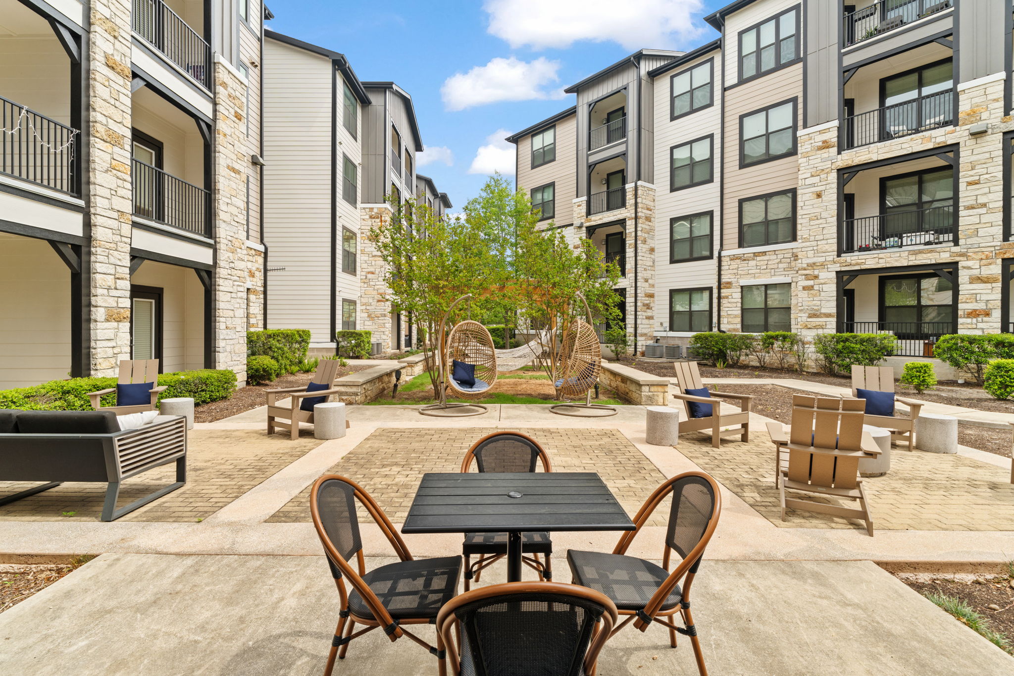 Apartment complex courtyard with outdoor dining tables, lounge chairs, hanging egg chairs, and surrounding residential buildings.