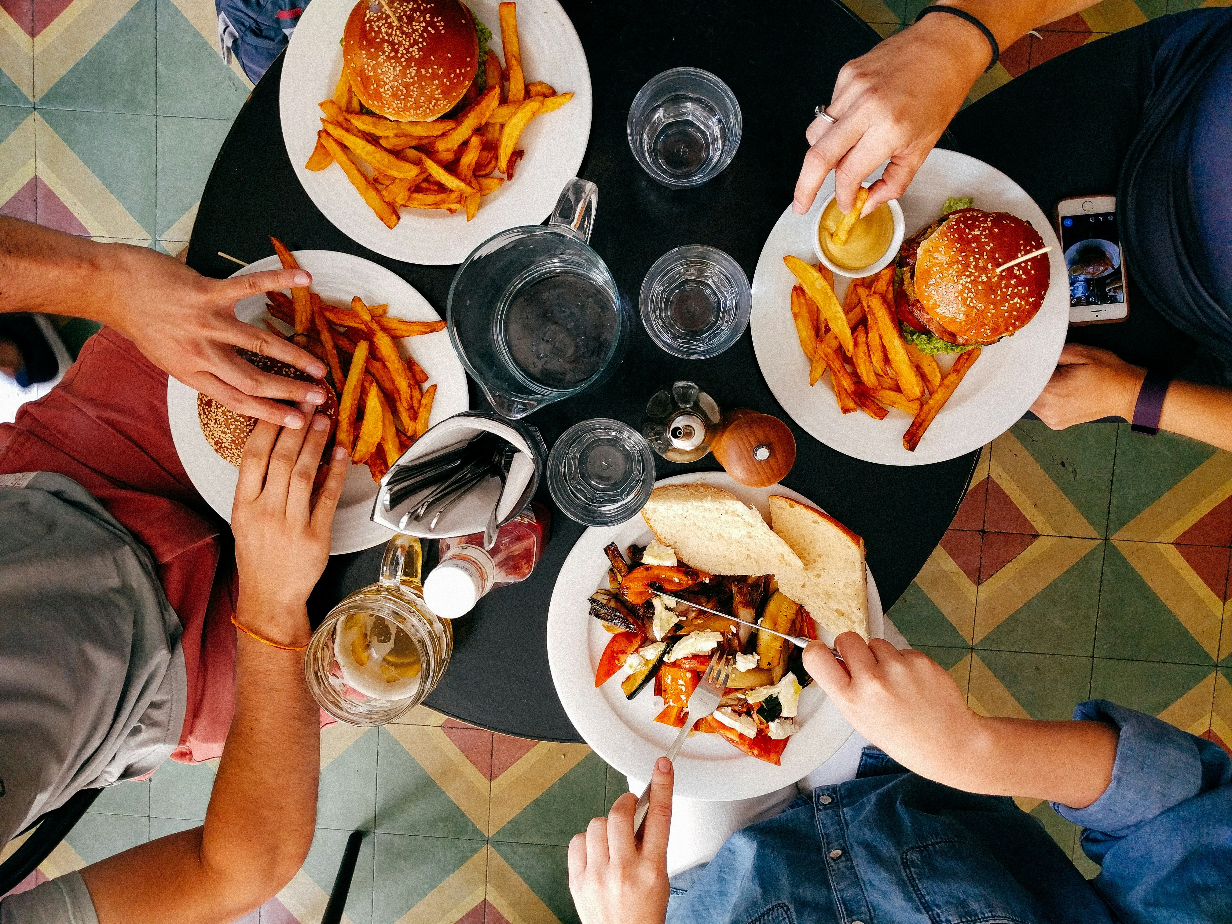 Overhead view of friends sharing burgers, fries, grilled vegetables, bread, and drinks around a restaurant table.