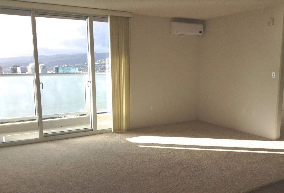 A room with a carpet floor, a window with a view of the city, and a sliding door.