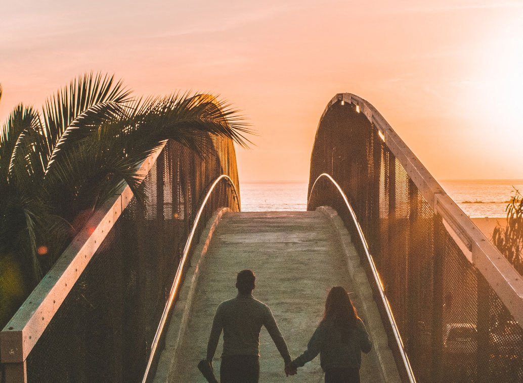 a man and woman walking down a bridge to the beach at sunset