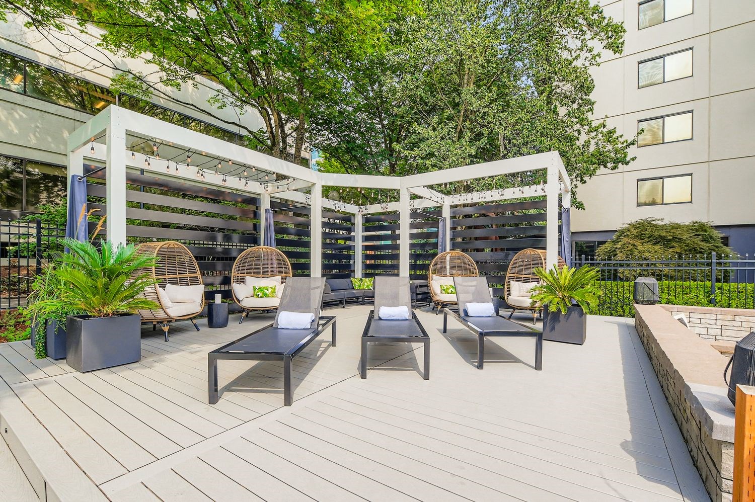 Modern Apartments in Downtown Portland, OR - Linc301 - Outdoor Lounge With Pergolas, Chaise Lounges, Hanging Egg Chairs, Potted Plants, Metal Fence, and Mature Trees.