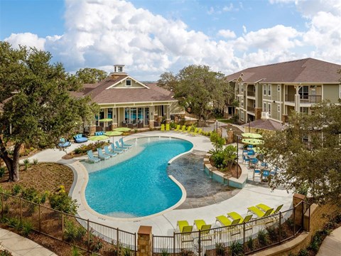 Aerial fenced in pool view with in and poolside seating surrounded by apartments, clubhouse and lush landscape