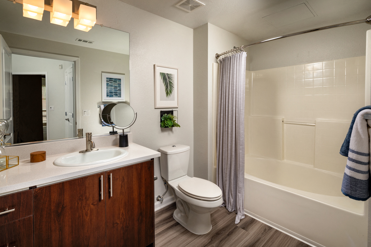 Apartments for Rent in La Mesa, CA - The District - A Bathroom with a Large Mirror, Wood Vanity, White Countertop, and a Shower/Tub.